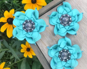 May include: Three turquoise leather flower hair clips with silver and crystal center embellishments. The flower petals are layered, creating a dimensional effect. The clips are arranged on a wooden surface, with yellow flowers and green foliage in the background.