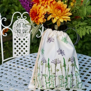 May include: A white cotton drawstring bag with a floral print design. The bag is sitting on a white metal table with a bouquet of orange and purple flowers in the background.