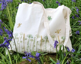 May include: Off-white canvas tote bag with a floral and bee design. The bag features a row of green and gold flowers along the bottom edge, with two gold bees and one green bee scattered across the front. The bag has long, off-white straps.