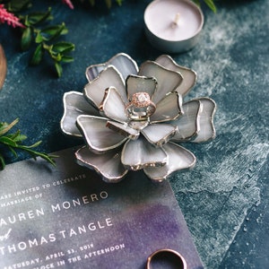 Blue stained-glass flower ring dish showcased with lavender candles and a wedding invitation in a romantic editorial flatlay. The soft color harmony highlights the engagement ring and complements invitation styling, engagement shoots, and refined bridal-party gifting moments.