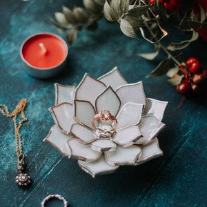 Handcrafted stained-glass flower ring dish with layered opaque white petals edged in silver solder, holding a rose-gold engagement ring on a deep teal surface, styled with a red candle, greenery, festive berries, and fine jewelry; elegant handmade jewelry holder for weddings and keepsakes.