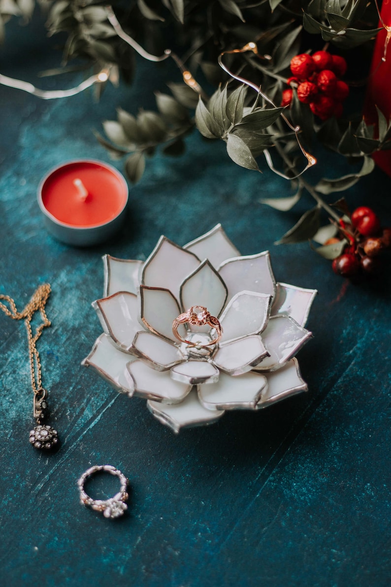 Handcrafted stained-glass flower ring dish with layered opaque white petals edged in silver solder, holding a rose-gold engagement ring on a deep teal surface, styled with a red candle, greenery, festive berries, and fine jewelry; elegant handmade jewelry holder for weddings and keepsakes.
