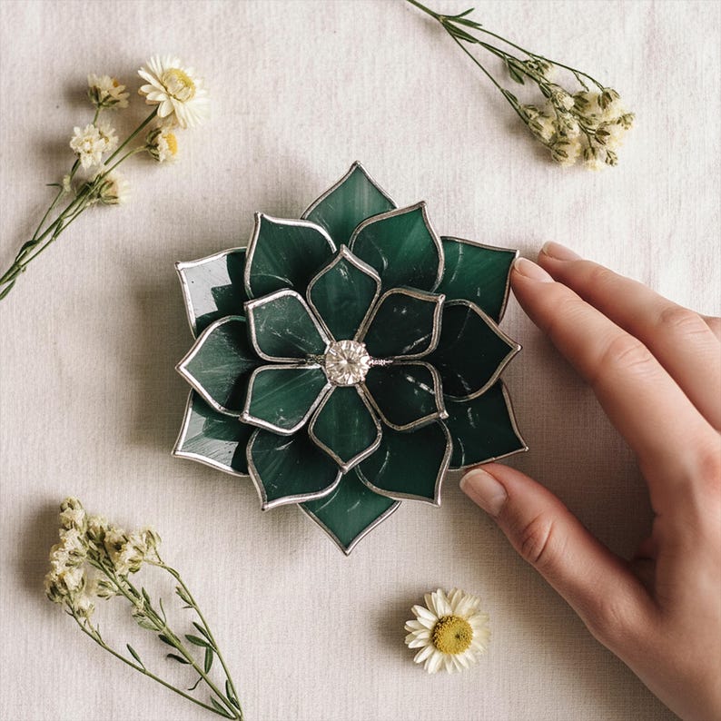 May include: A handcrafted, stained glass succulent in shades of emerald green with a silver-colored metal outline. The center of the succulent features a sparkling, clear crystal. The piece is displayed on a neutral background with dried flowers.