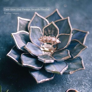 Puede incluir: Un portador de anillos en forma de flor de vidriera azul y plateada. Un anillo de oro con varias piedras transparentes se encuentra en el centro. El texto dice "Two-time Etsy Design Awards Finalist Wedding Category."
