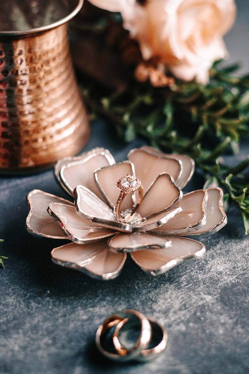 Handcrafted soft blush stained-glass flower ring dish holding a delicate engagement ring, styled with a copper vessel and botanical greenery. Soft-glam, vintage-romantic wedding aesthetics framed in an editorial flatlay, ideal for bridal morning details and elegant keepsake gifting.