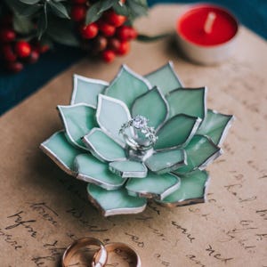 May include: A handcrafted, light green glass succulent ring holder with a silver ring featuring a large, clear gemstone. Two gold wedding bands are at the bottom. A red candle and berries are in the background. The ring holder is approximately 4 inches in diameter.