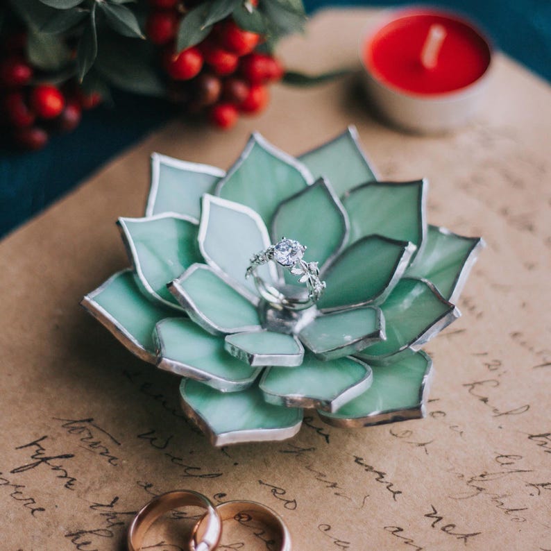 May include: A handcrafted, light green glass succulent ring holder with a silver ring featuring a large, clear gemstone. Two gold wedding bands are at the bottom. A red candle and berries are in the background. The ring holder is approximately 4 inches in diameter.