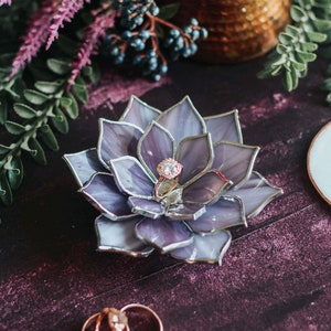 Handcrafted stained-glass flower ring dish with layered soft lavender and smoky gray petals edged in silver solder, holding a rose-gold engagement ring on a rich plum-toned surface, styled with greenery, dark berries, and a hammered copper vessel; elegant handmade jewelry holder for weddings and keepsakes.