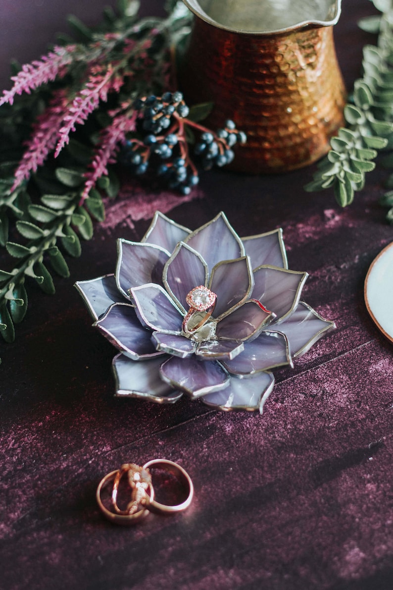 Handcrafted stained-glass flower ring dish with layered soft lavender and smoky gray petals edged in silver solder, holding a rose-gold engagement ring on a rich plum-toned surface, styled with greenery, dark berries, and a hammered copper vessel; elegant handmade jewelry holder for weddings and keepsakes.