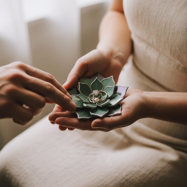 May include: A decorative, green and purple glass succulent ring holder with two gold rings with diamonds. The holder is being held by a person wearing a cream-colored dress.