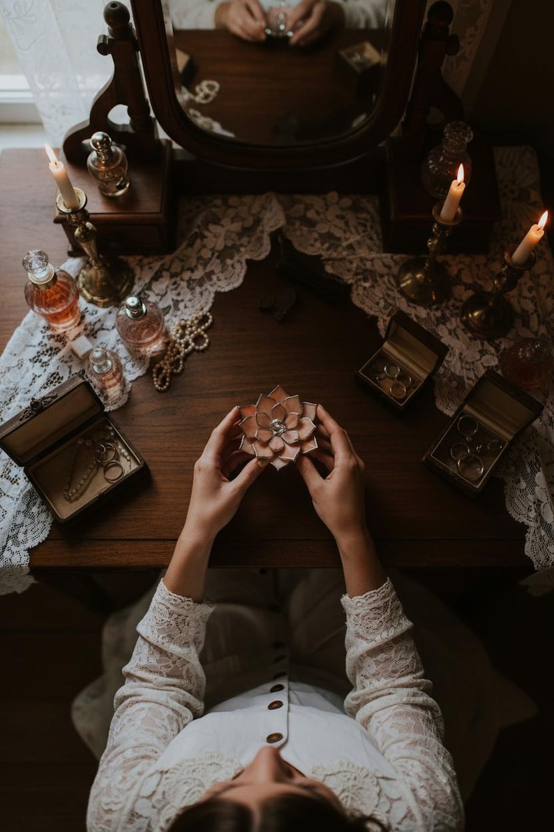 Puede incluir: Un tocador de estilo vintage con espejo, velas y frascos de perfume. Una joyero decorativo en forma de flor con una gema central se sostiene en las manos. Tambi&eacute;n hay otros joyeros y un collar de perlas sobre la mesa.