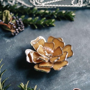 Amber stained-glass flower ring dish arranged among natural greenery on a deep teal surface. Warm amber tones suit autumn wedding trends, while the ring in the center adds a romantic editorial touch. Ideal for bridal party styling, fall flatlays, and unique ring-bearer alternatives.