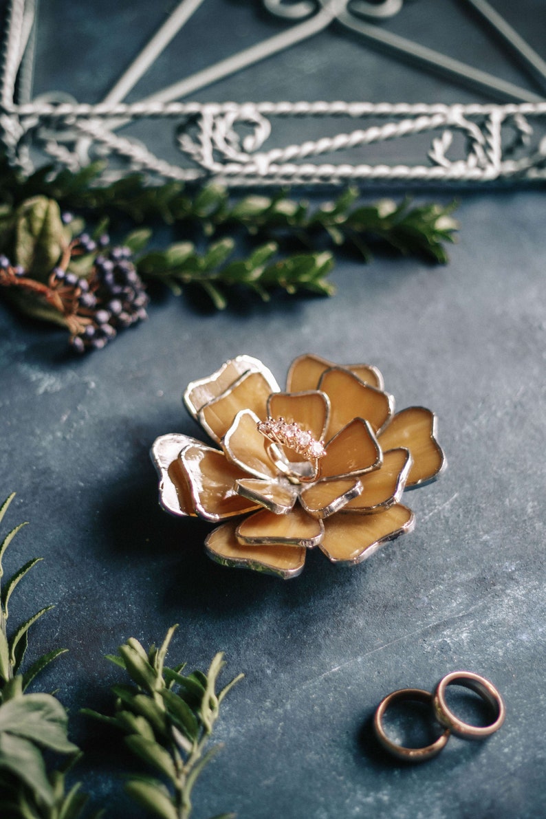 Amber stained-glass flower ring dish arranged among natural greenery on a deep teal surface. Warm amber tones suit autumn wedding trends, while the ring in the center adds a romantic editorial touch. Ideal for bridal party styling, fall flatlays, and unique ring-bearer alternatives.