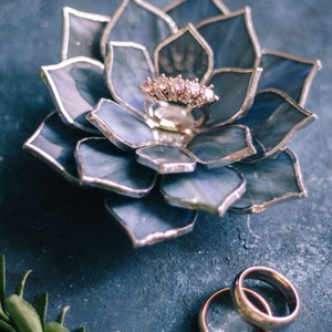 Handcrafted stained-glass flower ring dish with layered blue petals edged in silver solder, holding a gold engagement ring on a textured slate-blue surface, with gold wedding bands and subtle greenery nearby; elegant handmade jewelry holder for weddings and keepsakes.