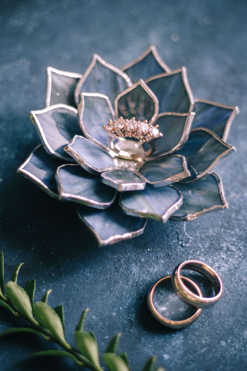 Handcrafted stained-glass flower ring dish with layered blue petals edged in silver solder, holding a gold engagement ring on a textured slate-blue surface, with gold wedding bands and subtle greenery nearby; elegant handmade jewelry holder for weddings and keepsakes.