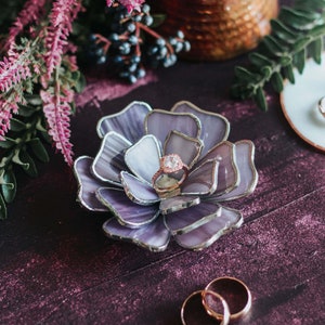 Lavender purple stained-glass floral ring dish styled as an elegant ring-bearer alternative, holding a rose-gold oval engagement ring in its layered petals. Set on a deep plum surface with soft botanical accents, copper details, and wedding bands nearby. A romantic, feminine heirloom piece for presenting rings with vintage glamour on the wedding day.