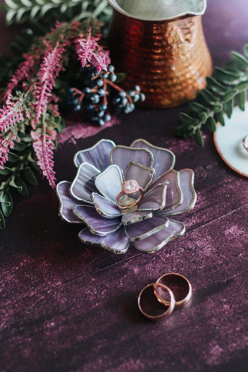 Lavender purple stained-glass floral ring dish styled as an elegant ring-bearer alternative, holding a rose-gold oval engagement ring in its layered petals. Set on a deep plum surface with soft botanical accents, copper details, and wedding bands nearby. A romantic, feminine heirloom piece for presenting rings with vintage glamour on the wedding day.