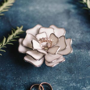 White stained-glass floral ring dish styled as an elegant ring-bearer alternative, featuring layered glass petals holding a rose-gold ring on a textured blue backdrop. Soft greenery frames the scene, with two wedding bands placed below. A refined, non-traditional option for carrying rings down the aisle, blending botanical charm with modern heirloom appeal.
