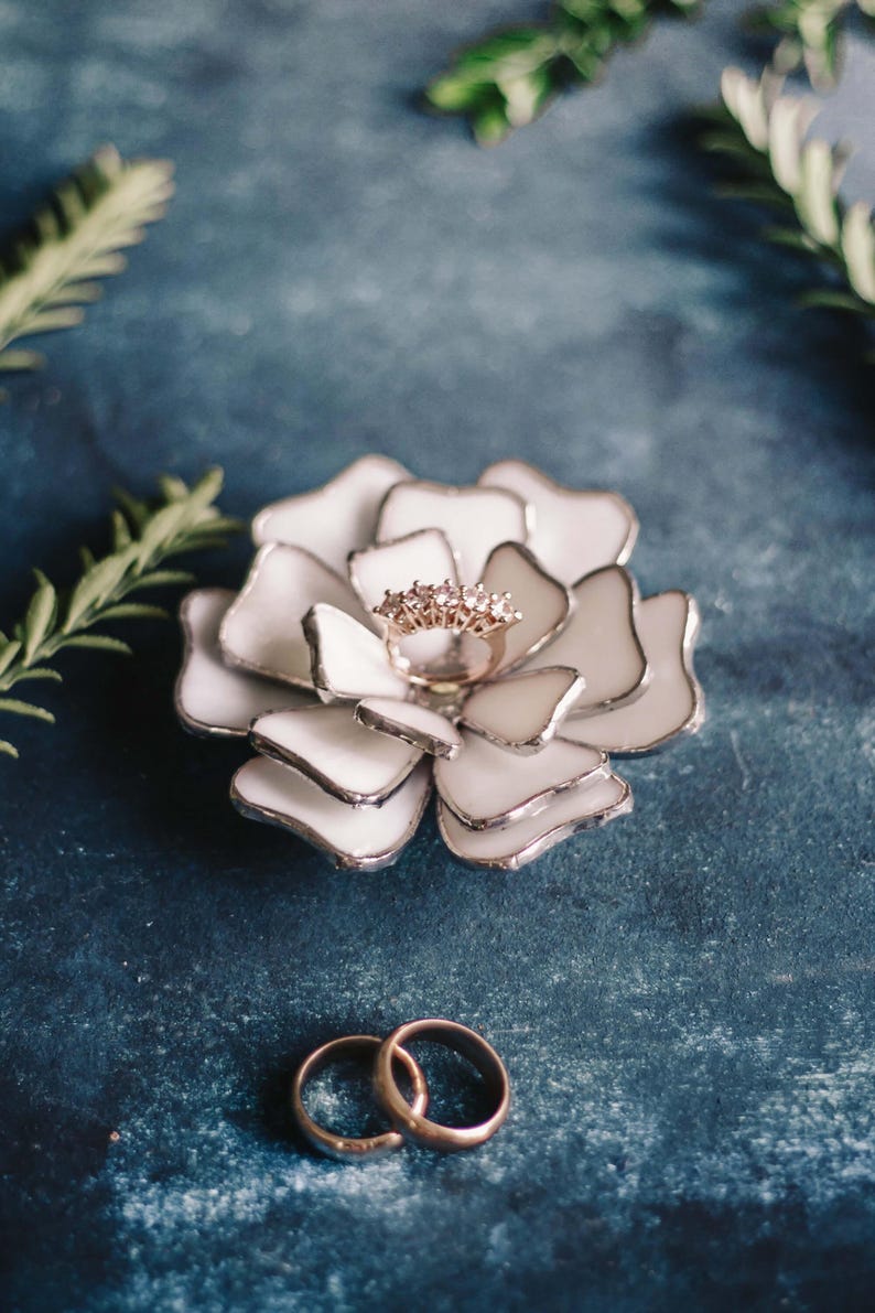 White stained-glass floral ring dish styled as an elegant ring-bearer alternative, featuring layered glass petals holding a rose-gold ring on a textured blue backdrop. Soft greenery frames the scene, with two wedding bands placed below. A refined, non-traditional option for carrying rings down the aisle, blending botanical charm with modern heirloom appeal.