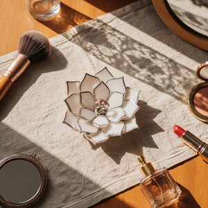 May include: A white and silver floral-shaped ring holder with a ring in the center, surrounded by makeup and accessories. A makeup brush, lipstick, perfume, and a compact mirror are arranged on a neutral-colored cloth.