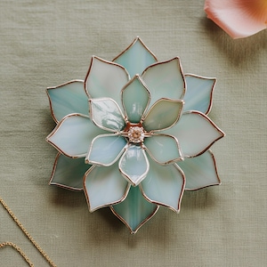 May include: A decorative stained glass flower in shades of light blue and white, with silver edging. A small, sparkling gem is set in the centre. The piece could be a brooch or ornament.