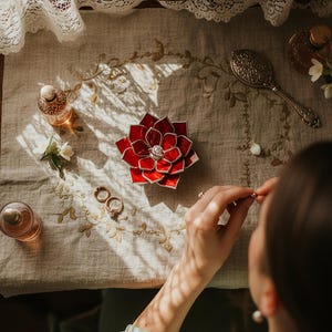 May include: A red, flower-shaped ring holder with a central diamond sits on a beige embroidered cloth. Two rings and perfume bottles are also on the table. A vintage mirror and hand mirror are in the background.