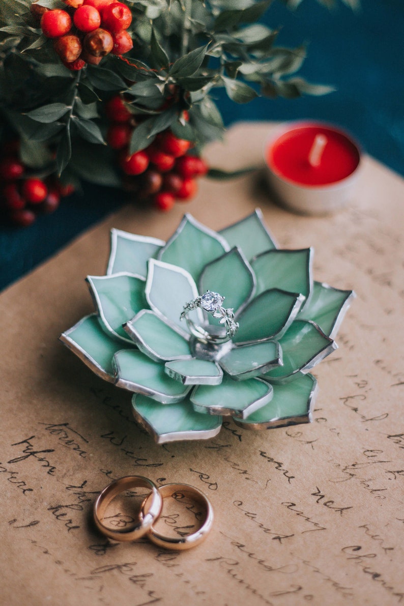 Handcrafted stained-glass succulent ring dish with layered green petals edged in silver solder, holding a diamond engagement ring on vintage handwritten paper, styled with red berries, a candle, and gold wedding bands; elegant handmade jewelry holder for weddings and keepsakes.