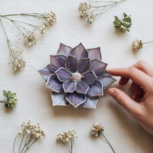 May include: A purple and silver stained glass flower-shaped ring holder. The ring holder has a silver border and a ring with a clear stone in the center. The background includes white flowers and greenery.