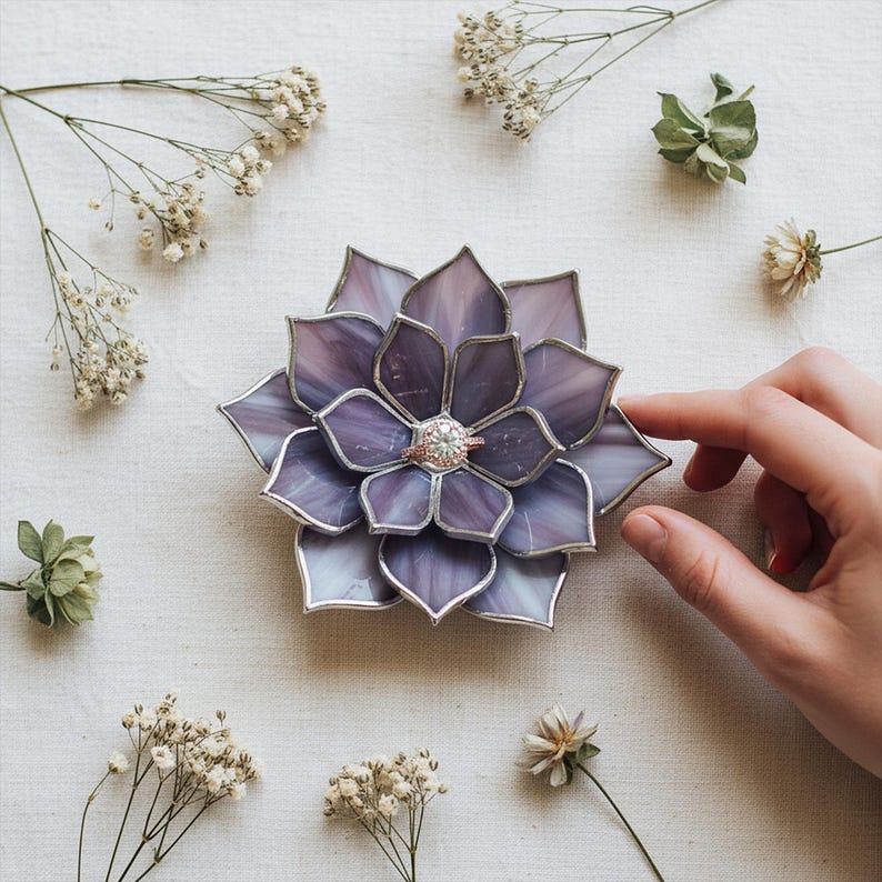 May include: A purple and silver stained glass flower-shaped ring holder. The ring holder has a silver border and a ring with a clear stone in the center. The background includes white flowers and greenery.