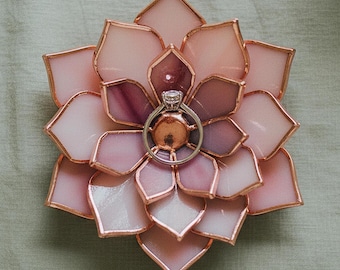 May include: A stained glass lotus flower dish in shades of pale pink and mauve with copper soldered edges. A silver-toned ring with a clear round gemstone is positioned in the center of the flower, resting on the flat copper base of the dish.