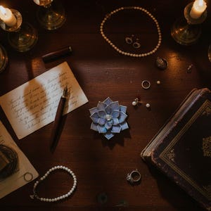 Puede incluir: Naturaleza muerta sobre una superficie de madera oscura con joyas: collar de perlas, pendientes, anillos y pulsera. Un broche en forma de flor azul y morada est&aacute; cerca de un libro antiguo y una carta manuscrita. Velas encendidas.