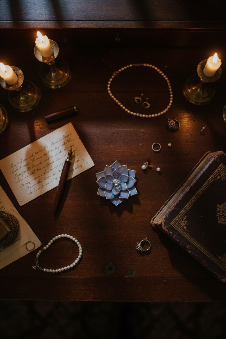 Puede incluir: Naturaleza muerta sobre una superficie de madera oscura con joyas: collar de perlas, pendientes, anillos y pulsera. Un broche en forma de flor azul y morada est&aacute; cerca de un libro antiguo y una carta manuscrita. Velas encendidas.