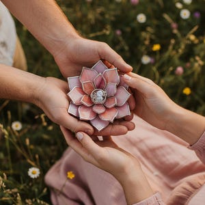 May include: A handcrafted, pink and peach-colored glass flower ring holder. The ring holder is held between two sets of hands, with a ring in the center. The background is a field of green grass and wildflowers.