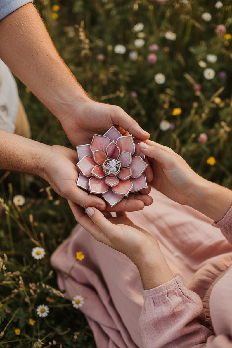 May include: A handcrafted, pink and peach-colored glass flower ring holder. The ring holder is held between two sets of hands, with a ring in the center. The background is a field of green grass and wildflowers.