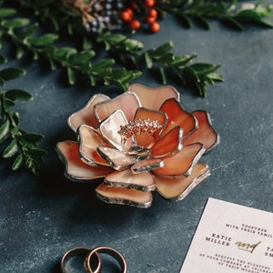 A peach-blush stained-glass flower ring dish with layered silver-edged petals holds a rose-gold engagement ring at its center. Styled on a deep-teal surface with green foliage, winter berries, gold wedding bands, and a wedding invitation, the scene creates a soft-glam, vintage bridal mood and highlights the dish as an elegant ring bearer and keepsake for weddings or engagements.