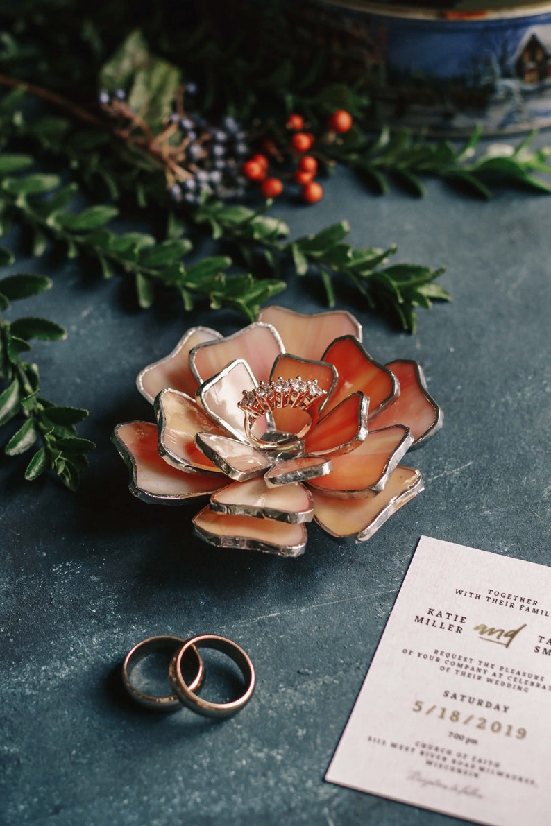 A peach-blush stained-glass flower ring dish with layered silver-edged petals holds a rose-gold engagement ring at its center. Styled on a deep-teal surface with green foliage, winter berries, gold wedding bands, and a wedding invitation, the scene creates a soft-glam, vintage bridal mood and highlights the dish as an elegant ring bearer and keepsake for weddings or engagements.