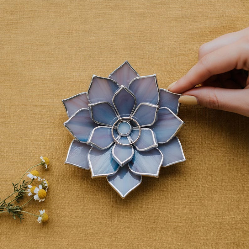 May include: A stained glass ring holder in the shape of a flower, with a silver ring in the center. The petals are a blend of blue and purple hues, outlined with silver solder. The holder sits on a yellow-gold surface.