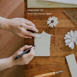 Puede incluir: Manos trabajando sobre un trozo de vidrio blanco, usando un rotulador negro para trazar un dise&ntilde;o. El texto "HANDCRAFTED from real stained glass" es visible. La superficie de madera tambi&eacute;n presenta un cortador de vidrio y recortes de flores de papel.