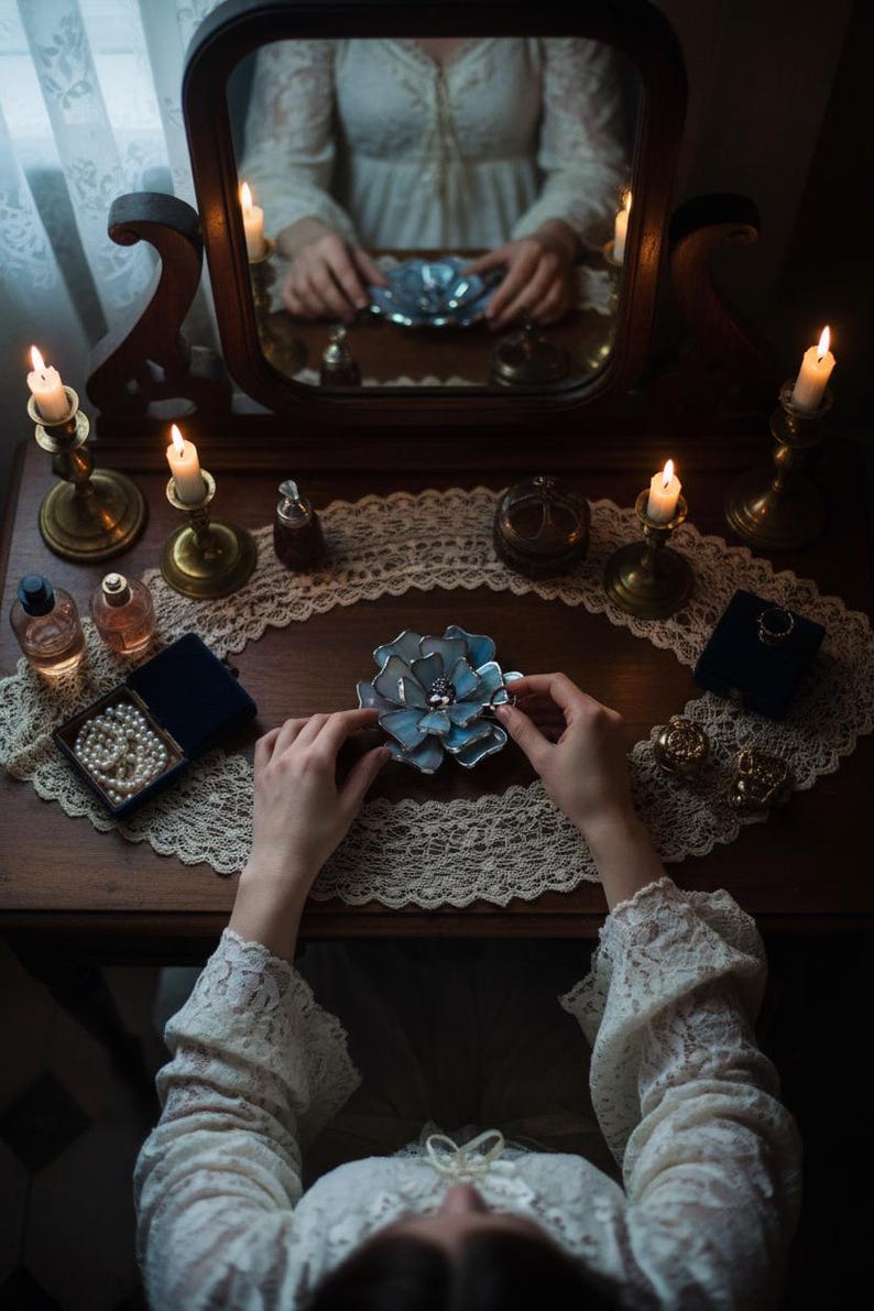 Puede incluir: Una escena de tocador vintage con espejo, velas y joyas. Las manos de una persona sostienen un objeto azul en forma de flor. Otros art&iacute;culos incluyen un collar de perlas en una caja azul, frascos de perfume y candelabros dorados. La escena est&aacute; iluminada con luz de velas.