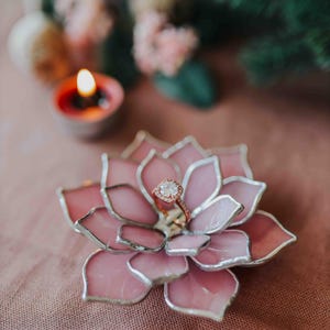May include: A pink, glass succulent ring holder with a silver outline, holding a rose gold ring with a diamond. A lit candle and greenery are in the background. The ring holder is on a pink surface.