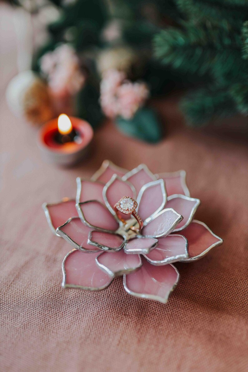 May include: A pink, glass succulent ring holder with a silver outline, holding a rose gold ring with a diamond. A lit candle and greenery are in the background. The ring holder is on a pink surface.