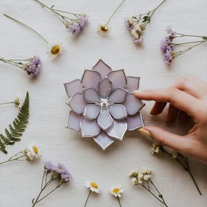 May include: A light purple, flower-shaped ring holder with a silver-colored metal outline. The ring holder is surrounded by small white and purple flowers and greenery. A hand is touching the ring holder.