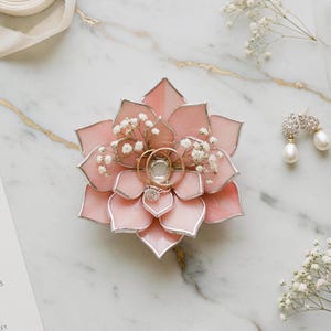 May include: A pink, flower-shaped ring holder with gold rings and a diamond ring. The holder is decorated with small white flowers. A pair of pearl earrings is also present on a marble surface.