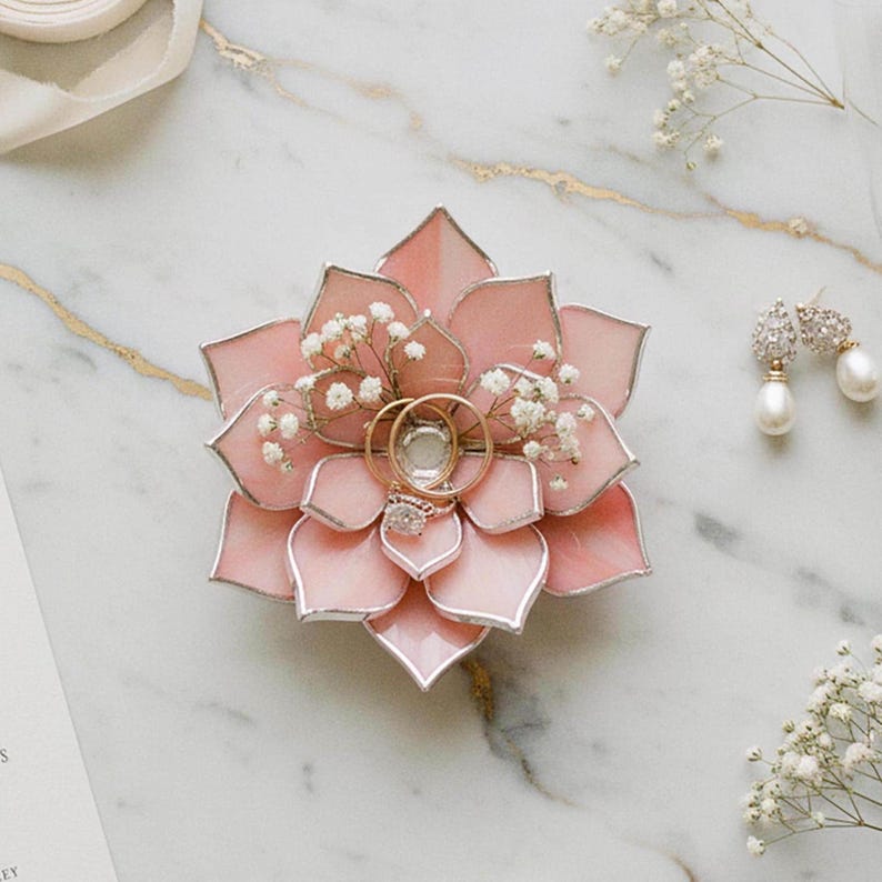 May include: A pink, flower-shaped ring holder with gold rings and a diamond ring. The holder is decorated with small white flowers. A pair of pearl earrings is also present on a marble surface.