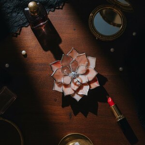 Puede incluir: Una flor decorativa de vidrieras con p&eacute;talos rosas y blancos, un borde plateado y una joya central. La imagen tambi&eacute;n incluye un frasco de perfume, un espejo compacto, un l&aacute;piz labial, una vela y perlas, todo dispuesto sobre una superficie de madera oscura.