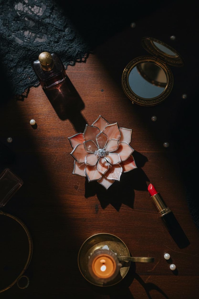 Puede incluir: Una flor decorativa de vidrieras con p&eacute;talos rosas y blancos, un borde plateado y una joya central. La imagen tambi&eacute;n incluye un frasco de perfume, un espejo compacto, un l&aacute;piz labial, una vela y perlas, todo dispuesto sobre una superficie de madera oscura.