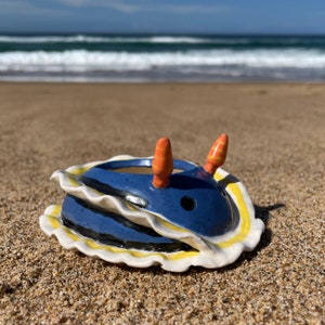 May include: A blue and white ceramic sea slug with orange and yellow accents. The sea slug is sitting on a sandy beach.