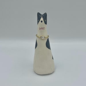 May include: A ceramic cat ornament in black and white, with a gold-coloured ring around its neck. The cat has a pink bow tie and black ears. The figurine stands on a white surface.