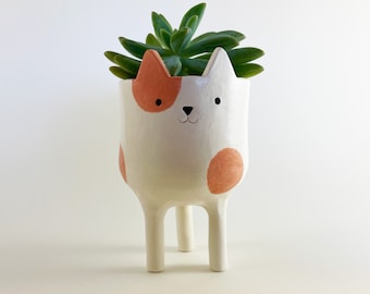 May include: A white ceramic planter shaped like a cat with three legs, featuring orange spots, black eyes, and a small nose. A green succulent plant is potted inside the container, which has pointed ears and a smooth, glossy finish.