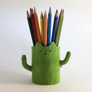 Op de afbeelding: Een groene keramische cactus potloodhouder met een schattig kattengezicht en uitgestrekte armen. De houder is gevuld met kleurpotloden.